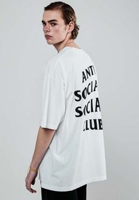 Młoda osoba z krótkimi włosami i kolczykiem, nosząca białą, oversize'ową koszulkę z nadrukiem "ANTI SOCIAL SOCIAL CLUB" na plecach.