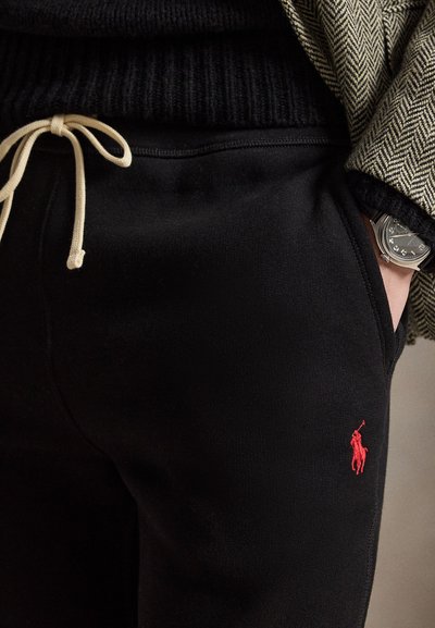 Pantaloni della tuta neri con coulisse beige e logo Polo rosso, indossati con giacca a spina di pesce e maglione nero, mano in tasca che mostra un orologio d'argento.