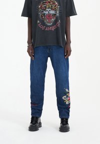 Camiseta gráfica negra con un diseño colorido de tigre; jeans de mezclilla azul de corte relajado con detalles bordados y un parche en la pierna.