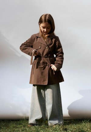 Bruine trenchcoat met dubbele knopen, riem en grote zakken, gecombineerd met wijdvallende, vervaagde denimspijkerbroeken. Grasachtergrond, neutrale achtergrond.