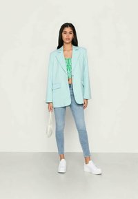 Blazer oversized azul claro, top curto verde estampado, jeans skinny azul claro e ténis brancos. Os acessórios incluem uma pequena bolsa branca.