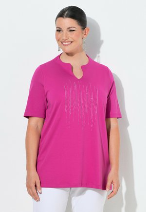 Mujer sonriente con cabello oscuro, con camiseta rosa de manga corta con diseño vertical de brillantes, lleva pendientes rectangulares de cristal, de pie frente a una pared blanca.