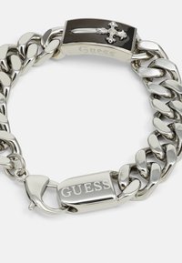 Stříbrný řetízek na zápěstí s obdélníkovým černým akcentem, který má design kříže. Na sponě je vyryto "Guess". Lesklý kovový povrch.
