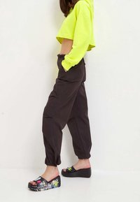 Neonowo zielona krótka bluza z kapturem, luźne czarne spodnie z kieszeniami oraz sandały na platformie z czarną podeszwą i kolorowymi wzorami graficznymi.