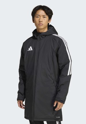 Junger Mann trägt eine lange schwarze Adidas-Kapuzensjacke mit weißen Streifen an den Ärmeln, steht vor einem schlichten hellen Hintergrund.