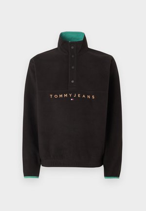 Čierny mikinový flís s vysokým golierom, štyrmi čiernymi cvokmi, zeleným vnútorným golierom a textom „TOMMY JEANS“ s logom na hrudi.
