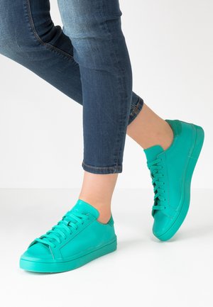 Osoba ubrana w niebieskie obcisłe dżinsy i jaskrawo-turkusowe sznurowane trampki, stojąca z jedną lekko uniesioną stopą.