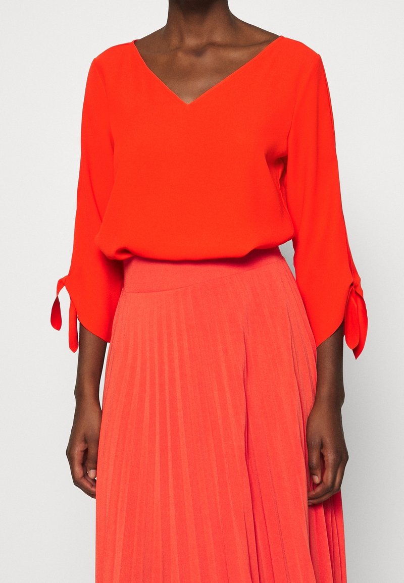 Blouse rouge à col en V avec manches trois-quarts et poignets noués, assortie à une jupe plissée dans une teinte plus claire de rouge.