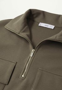 Jersey verde oliva con cuello de cremallera, con tejido texturizado y un bolsillo frontal. Herrajes de cremallera plateados que añaden detalle.