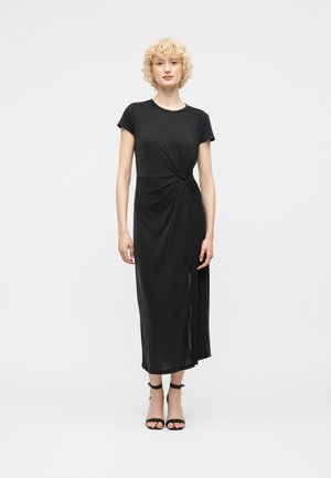 Kobieta z krótkimi, kręconymi blond włosami, ubrana w czarną, midi sukienkę z krótkim rękawem, z detalem w postaci węzła z boku i rozcięciem z przodu, zestawioną z czarnymi szpilkami z paskiem wokół kostki.