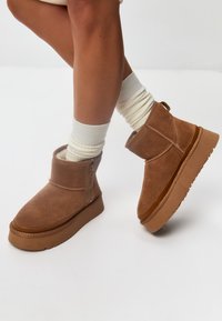 Braune Wildleder-Stiefeletten mit flacher Gummisohle, seitlichem Reißverschluss und genähten Akzenten. Getragen mit weißen gerippten Socken.