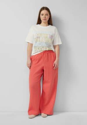 Młoda kobieta stoi przy szarej ścianie, ubrana w białą koszulkę z nadrukiem i luźne koralowe szerokie spodnie z beżowymi butami.