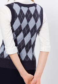 Marineblauwe en lichtblauwe argyle-gepatroneerde gebreide vest met een geribbelde zoom, gedragen over een witte blouse met lange mouwen. Uitzicht van de achterkant.