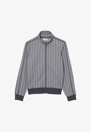 Chaqueta gris con cremallera, con patrón geométrico, puños y dobladillo oscuros, rayas a lo largo de las mangas y pequeño logotipo de cocodrilo verde en el pecho.