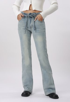 Kvinde iført lyseblå højttaljede denimjeans med papirpose-talje og bindebånd, sammen med sorte sko og en hvid cropped sweater.