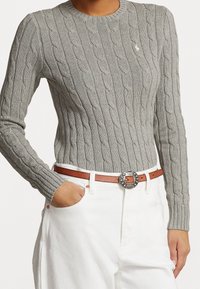 Personne portant un pull gris moulant en maille torsadée, rentré dans un pantalon blanc avec une ceinture en cuir marron ornée d'une boucle argentée décorative.
