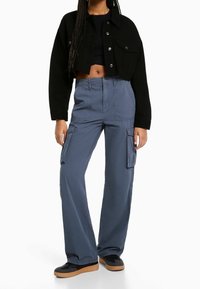 Svart kort jacka med knäppning, blå cargobyxor med sidofickor, och svarta sneakers med bruna sulor.