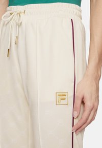 Crèmekleurige joggingsbroek met een textuurpatroon van Fila, elastische tailleband met een trekkoord en bordeauxrode zijstreep. Gouden logo-detail.