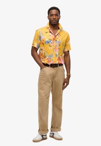 Chemise à manches courtes jaune à motifs floraux avec des accents roses et bleus, associée à un pantalon beige. Baskets blanches avec des rayures noires et vertes.