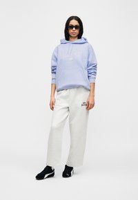 Ανοιχτό μωβ φούτερ με κουκούλα και μπροστινή τσέπη με λευκό γραφικό κείμενο, σε συνδυασμό με γκρι φαρδιά φόρμα με μαύρο λογότυπο, φορεμένο με μαύρα παπούτσια.