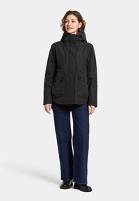 Svart vattentät jacka med huva, har två framfickor, dragkedjestängning och en något avslappnad passform. Buren med mörkblå vida jeans.