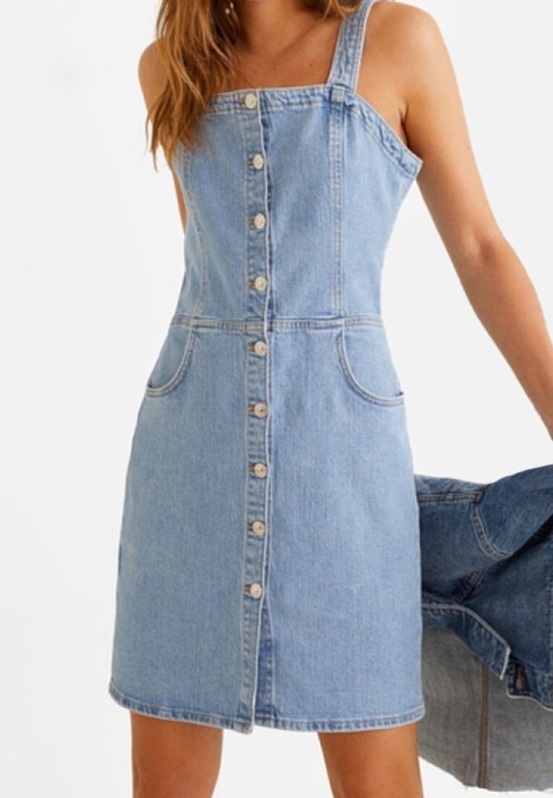 Femme portant une robe en jean bleu clair sans manches avec des boutons sur le devant et des poches, tenant une veste en jean dans une main.