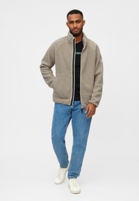 Fleece zip-up jakke i lys grå, med høj krave, to frontlommer og kontrastlynlås. Sammen med blå jeans og hvide sneakers.
