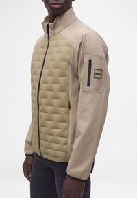 Homme portant une veste matelassée beige avec un col haut, une fermeture éclair noire et une poche zippée avec une étiquette "BOSS" sur la partie supérieure de la manche gauche.