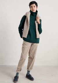 Mörkgrön stickad tröja med hög krage, beige kabelstickad halsduk, beige manchesterbyxor och mörka skor med mönstrade strumpor.