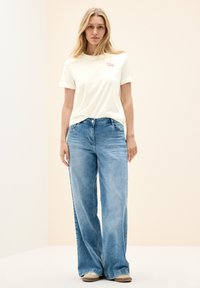T-shirt en coton jaune clair avec un logo rose, associée à un jean en denim bleu clair à coupe ample. Design simple, coupe décontractée, sans motifs notables.