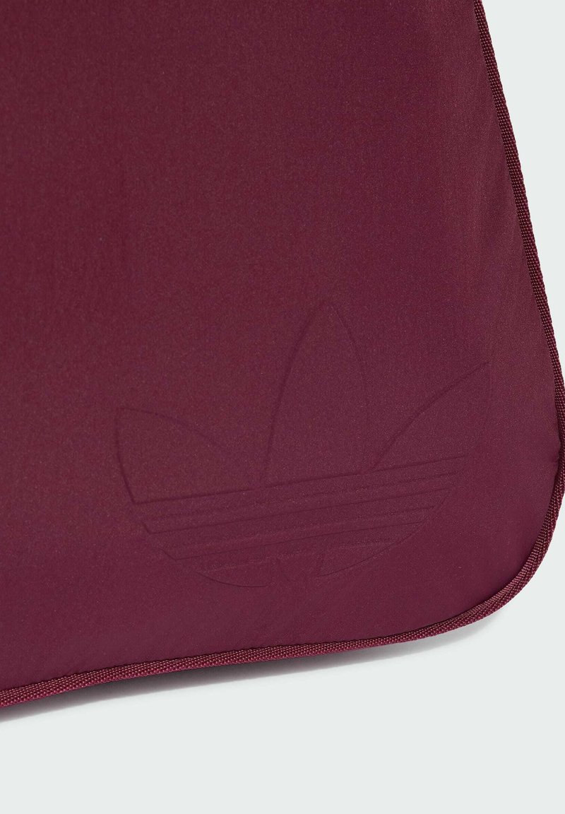 Tessuto color mogano con sottile logo Adidas trefoil in rilievo e piping abbinato color mogano lungo il bordo su uno sfondo chiaro.