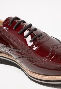 Chaussure brogue en cuir verni bourgogne avec des lacets noirs, des perforations détaillées et une semelle stratifiée avec des rayures beiges et noires.
