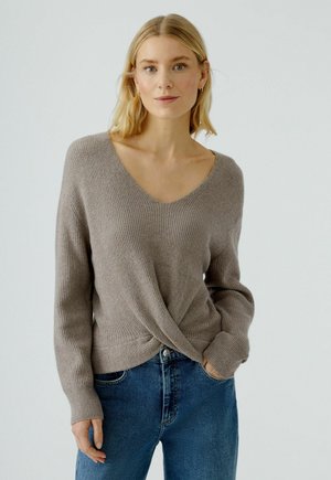 Hellbrauner, gerippter Strickpullover mit V-Ausschnitt und Twist-Design in der Taille, kombiniert mit blauen Jeans. Lässige lange Ärmel.