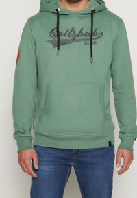 Grön hoodie med framficka, snoddar och broderad logotyp. Har en brun läderpatch på vänster arm. Mjuk tyggkänsla.