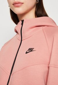 Rosa Nike Kapuzenpullover mit Reißverschluss, mit schwarzem Logo auf der Brust und kontrastierendem schwarzem Reißverschluss. Glattes Gewebe und sichtbares Kapuzendetail.