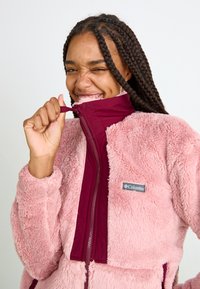 Fleecejakke i blush pink med en bordeaux lynlås. Har høj krave, tekstureret stof og et logo-patch på venstre bryst. Lynlås lukning.