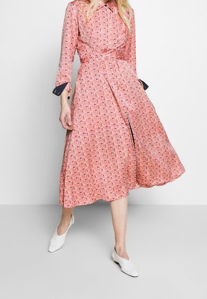 Vrouw draagt een roze midi-jurk met lange mouwen en patroon, met een kraag, gecombineerd met witte schoenen met lage hak, staand tegen een effen achtergrond.