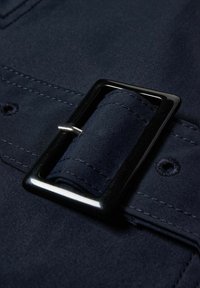 Granatowy pasek z materiału z prostokątną, czarną metalową klamrą i materiałową pętlą, widocznymi szwami i detalem w postaci guzika.