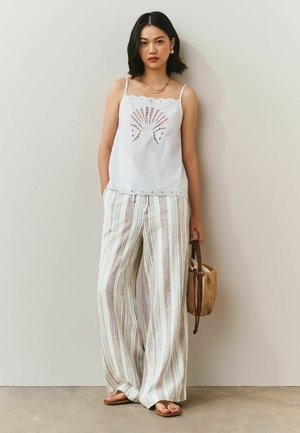 Kvinde iført hvid ærmeløs top med bølget kant og bælgudskæring samt brede, stribede bukser, der holder en flettet taske og bærer sandaler.