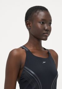 Débardeur de sport noir à fines bretelles, au design épuré, avec des accents gris texturés et un logo au niveau de la poitrine.