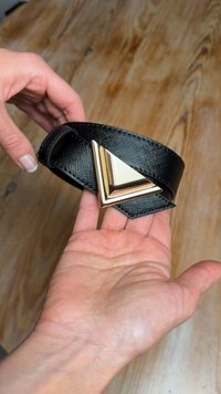 Črni teksturirani pas z zaponko v obliki trikotnika iz zlato obarvanega kovinskega materiala. Pas je držan v roki, kar poudarja njegov dizajn in obliko.