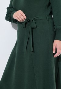 Abito verde scuro realizzato in materiale morbido, con cintura annodata in vita e maniche lunghe. Il tessuto ha una consistenza liscia con una leggera elasticità.