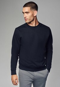 Marinblå crewneck sweatshirt i slätt tyg med ribbade ärmslut och nederkant, kombinerad med ljusgrå byxor. Enkelt design utan mönster.