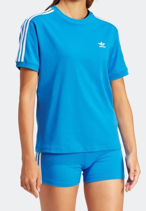 Femme portant un t-shirt bleu Adidas et un short assorti avec des bandes blanches sur les manches et les côtés du short, debout devant un fond uni.