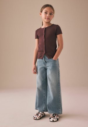 Mladá dívka s copánky stojí, má na sobě hnědý krátký rukáv s knoflíky, široké světle modré džíny a puntíkované nazouvací boty.