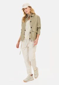 Oliven grøn letvægtsjakke med knapper, crème top, beige cargo-bukser og tan sneakers. Tilbehør inkluderer en lys cap og et mønstret tørklæde.