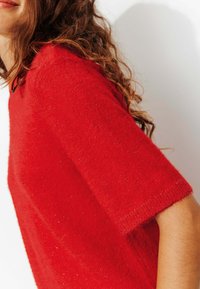 Pull rouge à manches courtes en tissu duveteux avec des paillettes subtiles. Caractérisé par une coupe décontractée et une surface douce et texturée avec un col rond.