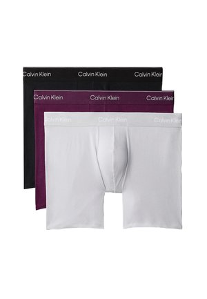 Junger Mann steht barfuß vor einfarbigem Hintergrund und trägt dunkelviolette Calvin Klein Boxershorts.