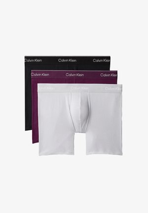 Jonge man staat blootsvoets tegen een effen achtergrond en draagt donkerpaarse Calvin Klein boxershorts.