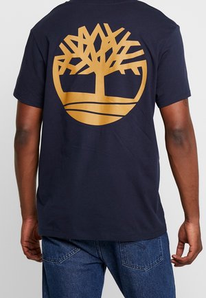 Person iført en mørkeblå T-shirt med stort gult trælogo på ryggen, kombineret med blå denimjeans.